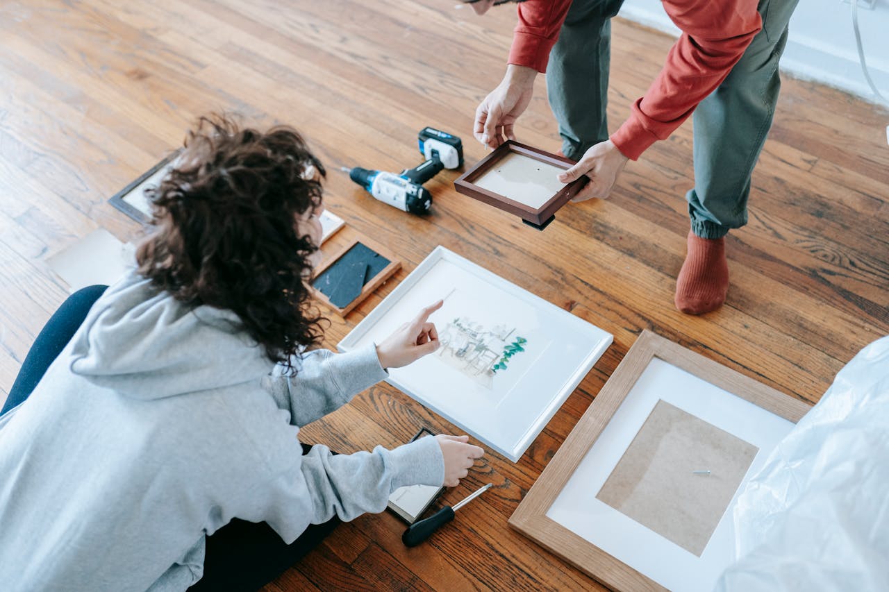 A couple organizing picture frames on a wooden floor, preparing for interior decoration or renovation.
