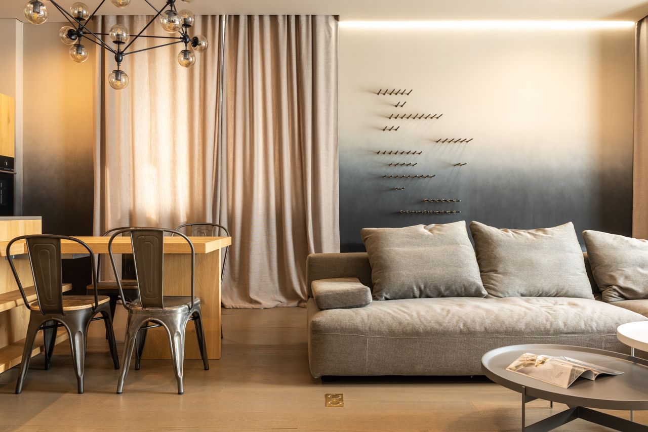 Comfortable couch with cushions and wooden table with chairs placed in modern living room with decorated wall and curtains at home
